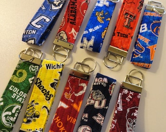 May include: A collection of ten colorful keychains with different college logos and team names. The keychains are made of fabric and have silver key rings. The logos include Colorado State, Wichita State, Virginia Tech, Alabama, Michigan, and others.