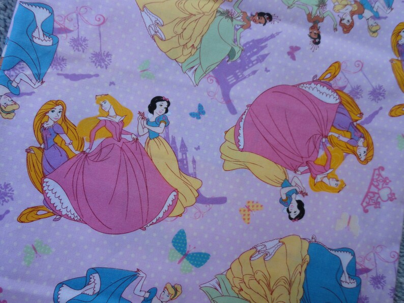 Pu&ograve; includere: Un tessuto rosa e viola con un motivo ripetitivo di principesse Disney. Le principesse indossano i loro abiti caratteristici e sono circondate da farfalle e castelli.