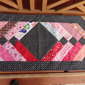 May include: A black and white quilted table runner with a diamond pattern in the center. The runner is made of various pink and red fabrics with different patterns. The runner is approximately 18 inches wide and 36 inches long.