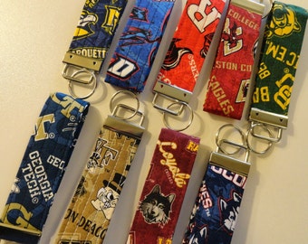 May include: A collection of nine fabric key fobs featuring various collegiate sports team logos and colors. Each rectangular strap is finished with a silver-toned metal clamp and a circular split ring for attaching keys, arranged in two rows.