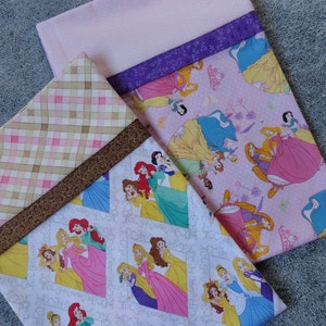 Pu&ograve; includere: Tre pannelli di tessuto a tema principessa. Il pannello superiore &egrave; rosa con un bordo viola e presenta varie principesse Disney nei loro abiti. Il pannello centrale &egrave; un plaid marrone e rosa con un bordo di nastro marrone. Il pannello inferiore &egrave; bianco con un motivo grigio e presenta varie principesse Disney nei loro abiti.