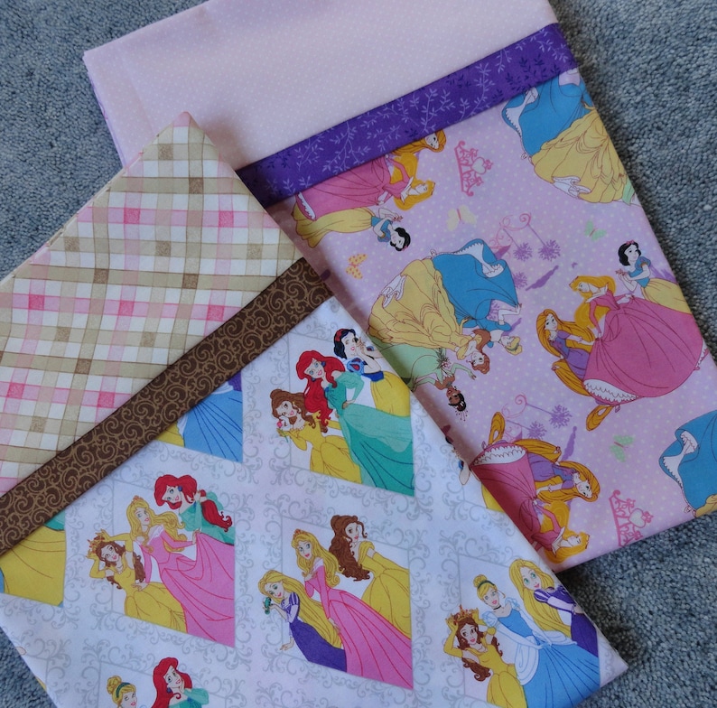 Pu&ograve; includere: Tre pannelli di tessuto a tema principessa. Il pannello superiore &egrave; rosa con un bordo viola e presenta varie principesse Disney nei loro abiti. Il pannello centrale &egrave; un plaid marrone e rosa con un bordo di nastro marrone. Il pannello inferiore &egrave; bianco con un motivo grigio e presenta varie principesse Disney nei loro abiti.