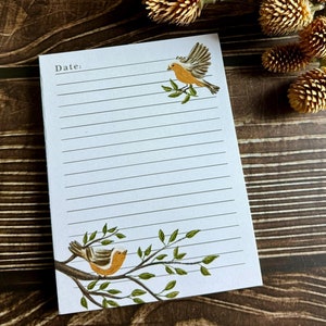 May include: A white notepad with lined paper and a bird illustration on the front. The notepad has the word "Date:" printed at the top. The notepad is on a brown wooden surface.
