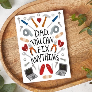 May include: A greeting card with the text "Dad, You Can Fix Anything" surrounded by illustrations of tools, bandages, and a broken heart. The card is on a wooden tray with eucalyptus leaves.