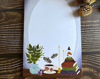 Op de afbeelding: Een witte kaart met een grijs-witte achtergrond. De kaart heeft een grote ovale vorm in het midden. De ovaal wordt omringd door een theekopje met schotel, een vaas met groene bladeren, twee motten, drie kaarsen, een glazen stolp met paddenstoelen en een stapel boeken. De boeken hebben gouden accenten en de tekst "The Book of Shadows" is zichtbaar op het bovenste boek.