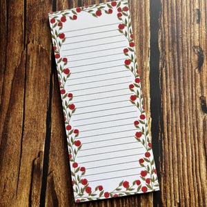 Könnte beinhalten: Ein rechteckiger Notizblock mit einem Rand aus roten Rosen und grünen Blättern. Das linierte Papier ist weiß mit schwarzen Linien. Auf einer Holzoberfläche platziert.
