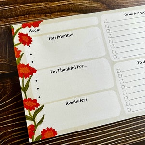 Könnte beinhalten: Ein Wochenplaner mit floralem Rand. Der Planer hat Abschnitte für "Woche:", "Top Prioritäten", "Ich bin dankbar für..." und "Erinnerungen". Es gibt auch Abschnitte für "Zu erledigen für die Arbeit" und "Zu erledigen".