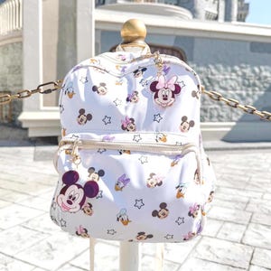 May include: White backpack featuring a repeating pattern of Mickey Mouse, Minnie Mouse, Donald Duck, and stars. The backpack has a gold zipper and a Minnie Mouse charm. The background includes a castle.