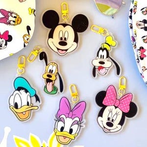May include: A collection of Disney-themed accessories, including keychains featuring Mickey Mouse, Donald Duck, Goofy, and Minnie Mouse. The image also shows a pair of sunglasses and two patterned bags with Disney character prints. The items are arranged on a light blue surface.