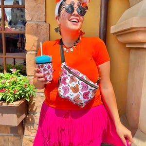 May include: A bright orange short-sleeved t-shirt, a vibrant pink tiered skirt, and a white fanny pack featuring a repeating pattern of orange and pink pumpkin-shaped mouse ears and black bats. The outfit includes white mouse ear headbands.