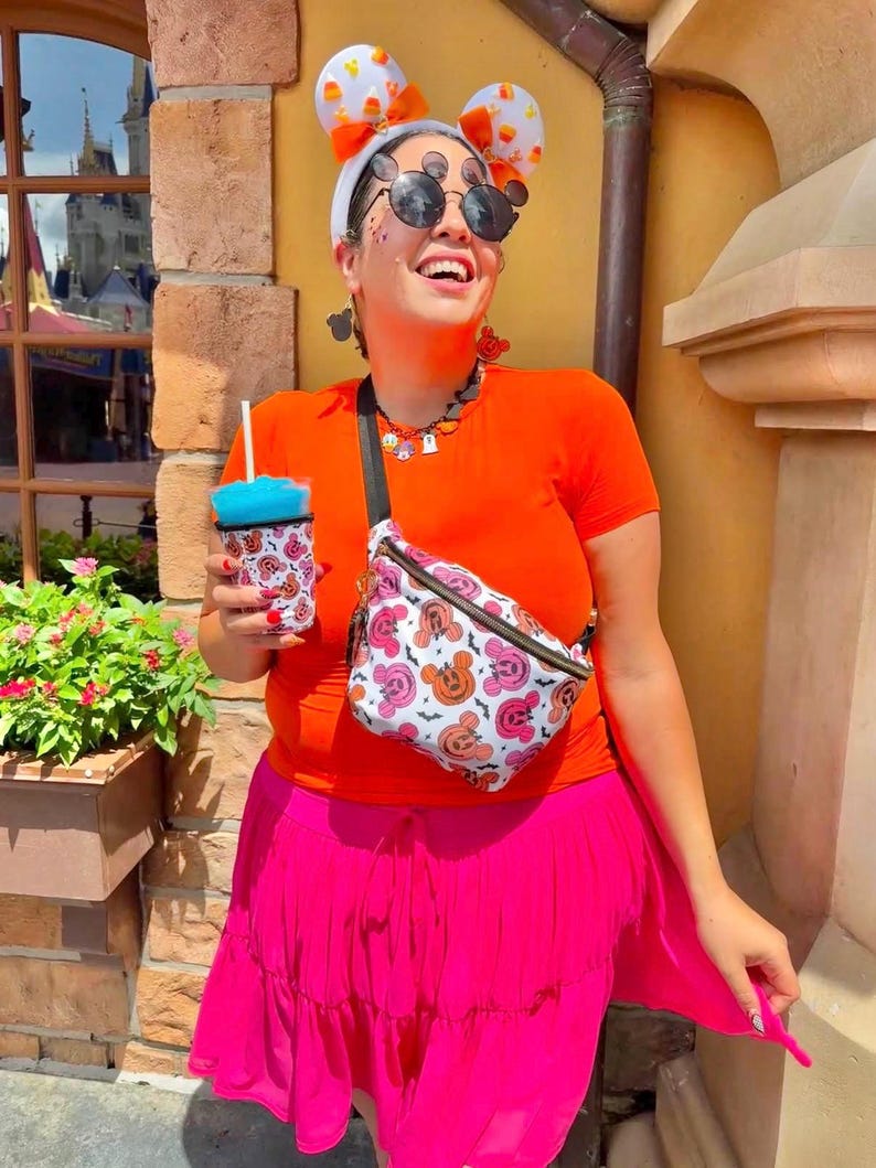 May include: A bright orange short-sleeved t-shirt, a vibrant pink tiered skirt, and a white fanny pack featuring a repeating pattern of orange and pink pumpkin-shaped mouse ears and black bats. The outfit includes white mouse ear headbands.