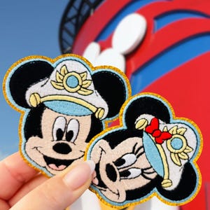 May include: Two embroidered patches featuring Mickey and Minnie Mouse. Mickey wears a white captain's hat with a gold trim and a blue band. Minnie has a red bow and a matching hat. Both have gold borders.