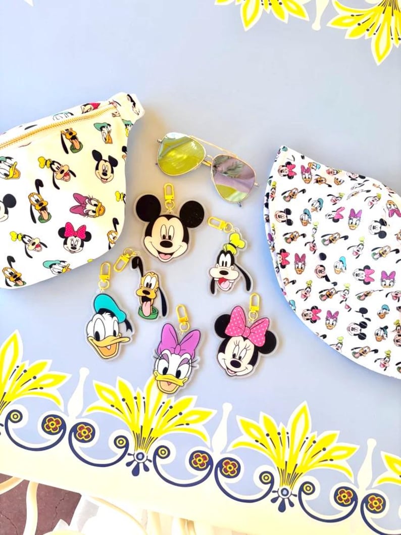May include: A collection of Disney-themed accessories, including keychains featuring Mickey Mouse, Donald Duck, Goofy, and Minnie Mouse. The image also shows a pair of sunglasses and two patterned bags with Disney character prints. The items are arranged on a light blue surface.