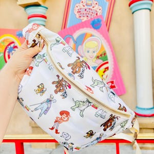 May include: White fanny pack with a gold zipper and a cream-colored strap. The bag is covered in colorful cartoon character illustrations. The characters include Woody, Buzz Lightyear, and other characters from the Toy Story franchise.