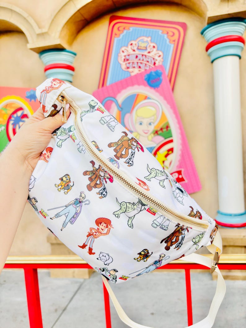 May include: White fanny pack with a gold zipper and a cream-colored strap. The bag is covered in colorful cartoon character illustrations. The characters include Woody, Buzz Lightyear, and other characters from the Toy Story franchise.