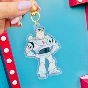 May include: A clear acrylic keychain featuring Buzz Lightyear from Toy Story. The keychain has a gold-colored clasp and a cartoon image of Buzz in his spacesuit. The background is a light blue color.
