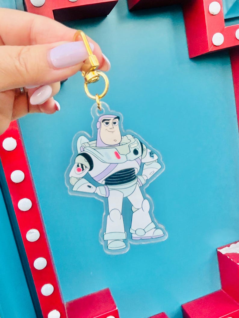 May include: A clear acrylic keychain featuring Buzz Lightyear from Toy Story. The keychain has a gold-colored clasp and a cartoon image of Buzz in his spacesuit. The background is a light blue color.