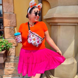 May include: A bright orange short-sleeved t-shirt, a vibrant hot pink tiered skirt, and a white fanny pack featuring a colorful Mickey Mouse pattern. The outfit includes white mouse-ear headbands with orange accents and a matching patterned cup sleeve.