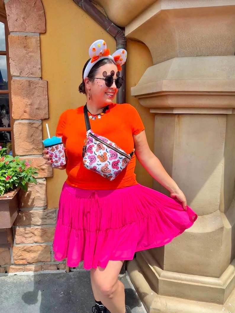 May include: A bright orange short-sleeved t-shirt, a vibrant hot pink tiered skirt, and a white fanny pack featuring a colorful Mickey Mouse pattern. The outfit includes white mouse-ear headbands with orange accents and a matching patterned cup sleeve.