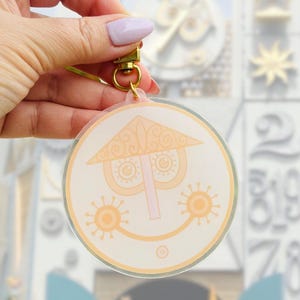May include: A clear, round keychain featuring a smiling face design with a hat and sun-like accents in shades of peach and orange. The keychain has a gold-colored clasp and a light green strap.
