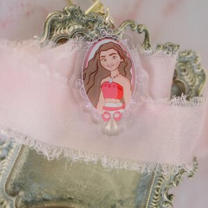 May include: A clear acrylic keychain featuring a cartoon illustration of a woman in a red top and brown hair. The keychain is set against a pink ribbon and a decorative, ornate metal tray.