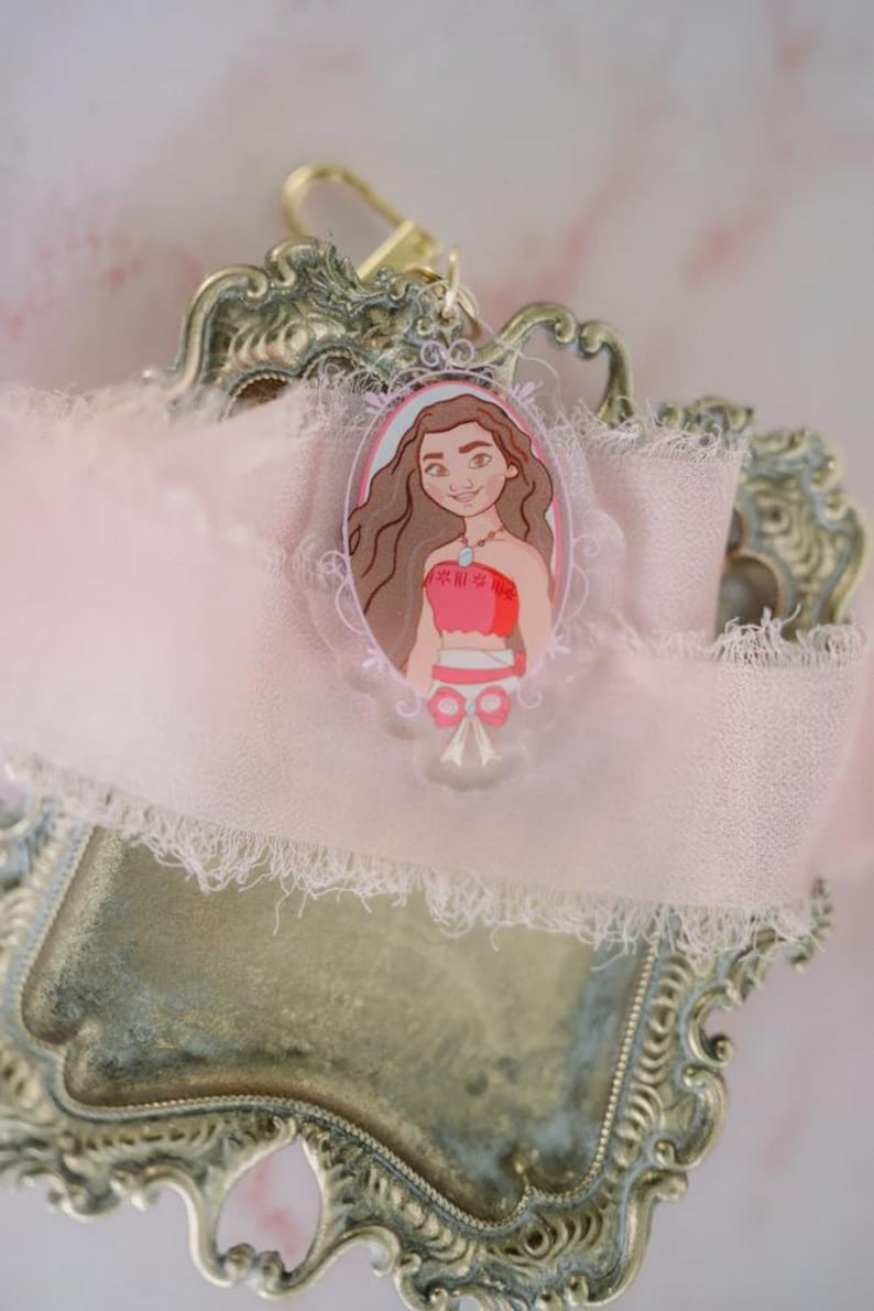 May include: A clear acrylic keychain featuring a cartoon illustration of a woman in a red top and brown hair. The keychain is set against a pink ribbon and a decorative, ornate metal tray.