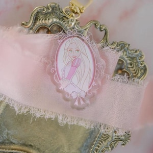 May include: A Rapunzel-themed keychain with a gold clasp. The acrylic charm features a pink and white illustration of Rapunzel in an oval frame. The charm is attached to a pink ribbon and rests on a decorative, ornate gold-colored tray.