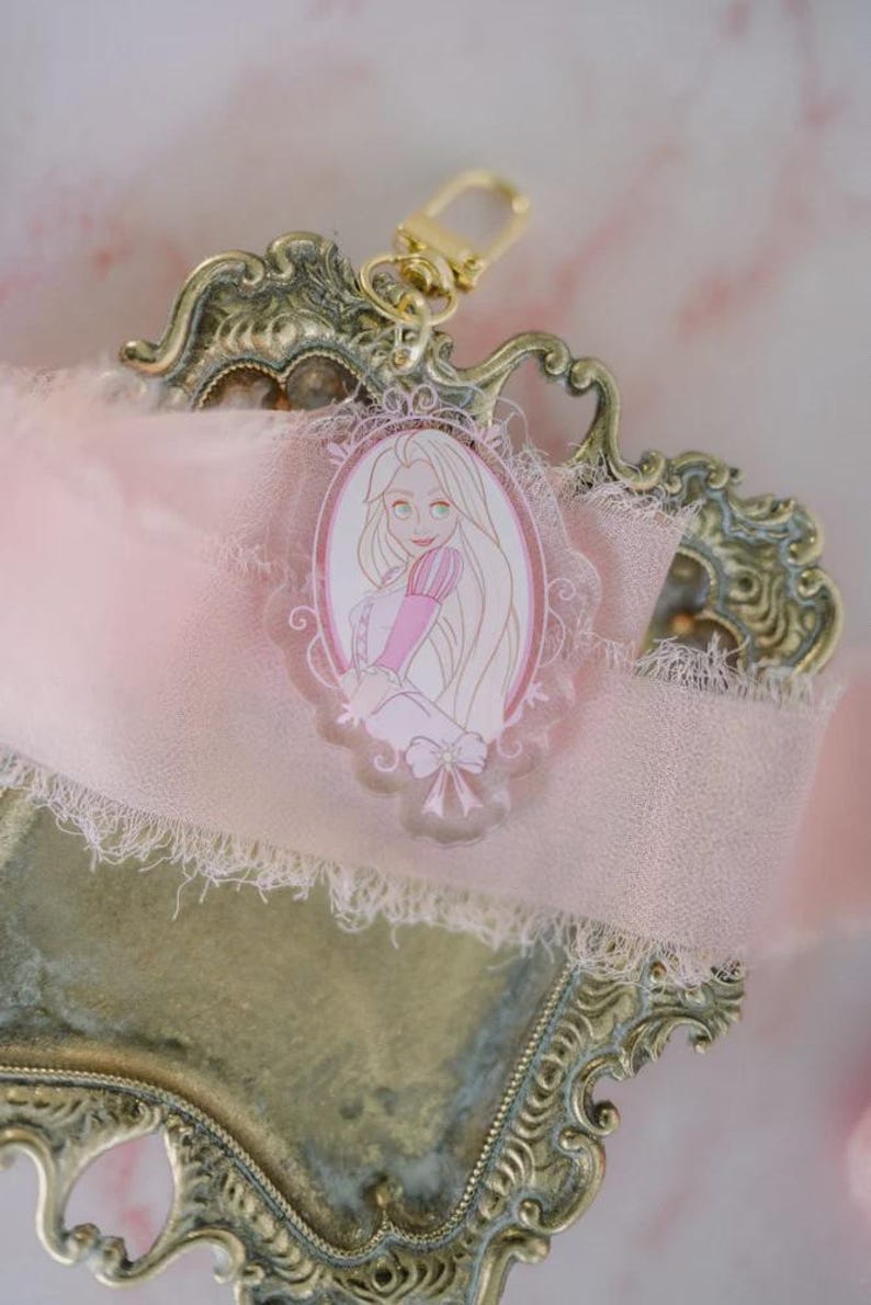 May include: A Rapunzel-themed keychain with a gold clasp. The acrylic charm features a pink and white illustration of Rapunzel in an oval frame. The charm is attached to a pink ribbon and rests on a decorative, ornate gold-colored tray.
