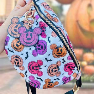 May include: A white fanny pack featuring a repeating pattern of orange and purple pumpkin-shaped mouse heads and small black bats. The bag includes a gold zipper and two matching pumpkin-shaped mouse head charms attached to the zipper pull.