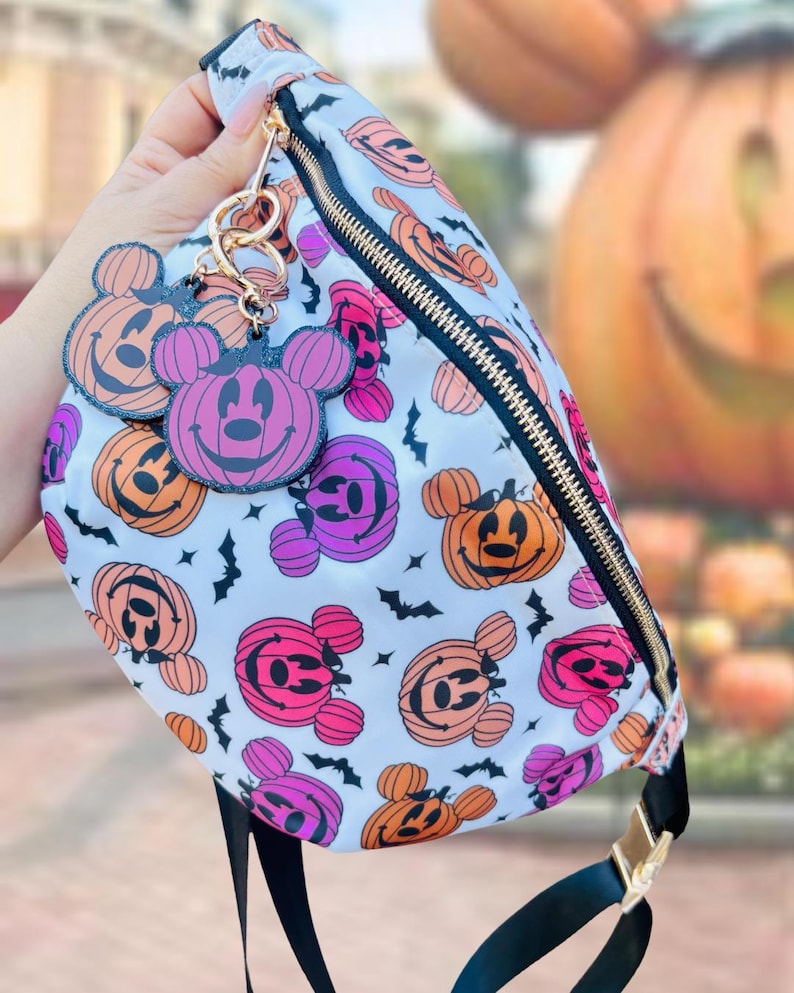 May include: A white fanny pack featuring a repeating pattern of orange and purple pumpkin-shaped mouse heads and small black bats. The bag includes a gold zipper and two matching pumpkin-shaped mouse head charms attached to the zipper pull.