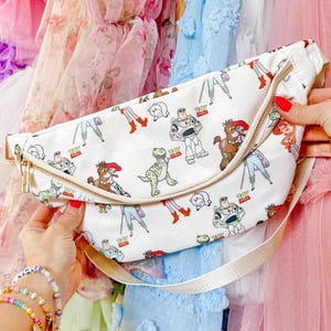 May include: White fanny pack with a gold zipper and a cream-colored strap. The bag is patterned with cartoon characters, including a dinosaur, a space ranger, and a cowboy. The bag is held by a person.