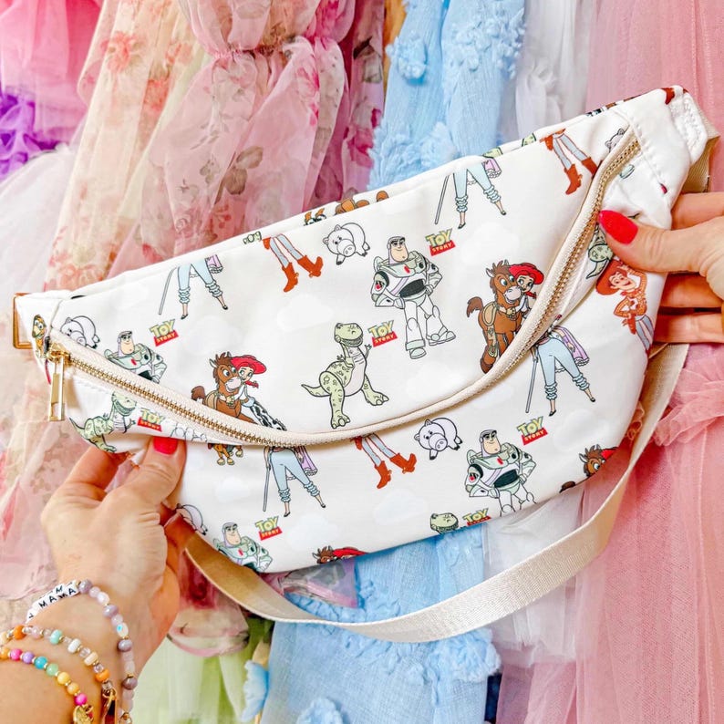 May include: White fanny pack with a gold zipper and a cream-colored strap. The bag is patterned with cartoon characters, including a dinosaur, a space ranger, and a cowboy. The bag is held by a person.