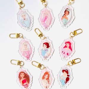 May include: A collection of acrylic keychains featuring various princess illustrations within ornate, pink-trimmed oval frames. Each keychain has a gold-colored clasp. The princesses are depicted in their signature outfits and hairstyles.