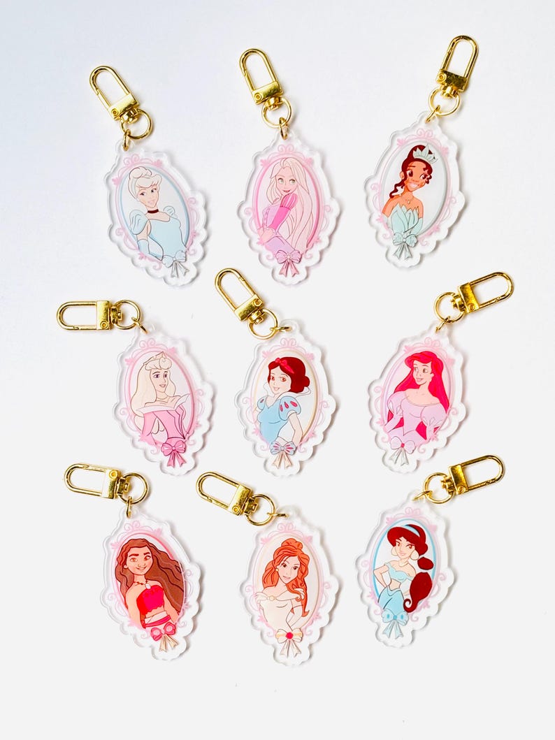 May include: A collection of acrylic keychains featuring various princess illustrations within ornate, pink-trimmed oval frames. Each keychain has a gold-colored clasp. The princesses are depicted in their signature outfits and hairstyles.