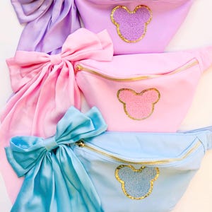May include: Three fanny packs with a bow and a Mickey Mouse head design. The fanny packs are in shades of purple, pink, and blue.