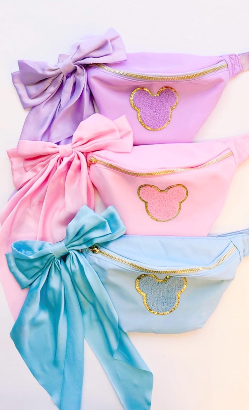 May include: Three fanny packs with a bow and a Mickey Mouse head design. The fanny packs are in shades of purple, pink, and blue.