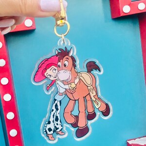 May include: A clear acrylic keychain featuring a cartoon image of Jessie and Bullseye from Toy Story. Jessie is wearing a pink hat and cowgirl outfit, hugging Bullseye, a brown horse. The keychain has a gold-colored clasp.