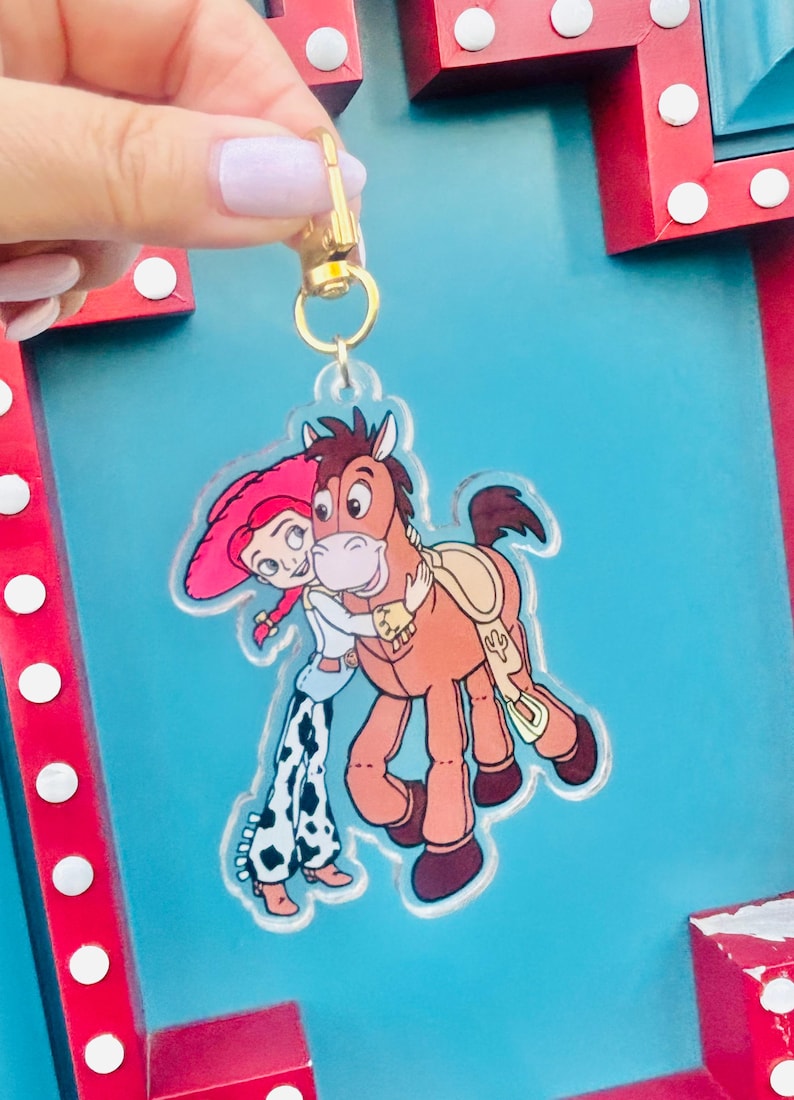 May include: A clear acrylic keychain featuring a cartoon image of Jessie and Bullseye from Toy Story. Jessie is wearing a pink hat and cowgirl outfit, hugging Bullseye, a brown horse. The keychain has a gold-colored clasp.
