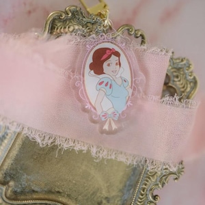 May include: A Snow White-themed keychain charm. The charm features an oval portrait of Snow White in a blue and white dress, with a pink bow. The charm is attached to a pink ribbon with a gold-colored clasp, set on a decorative gold-toned tray.