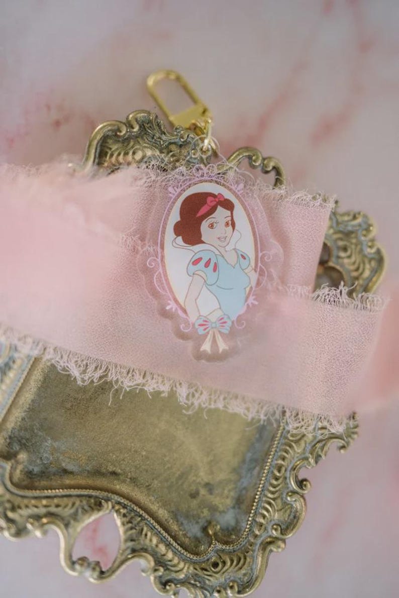 May include: A Snow White-themed keychain charm. The charm features an oval portrait of Snow White in a blue and white dress, with a pink bow. The charm is attached to a pink ribbon with a gold-colored clasp, set on a decorative gold-toned tray.