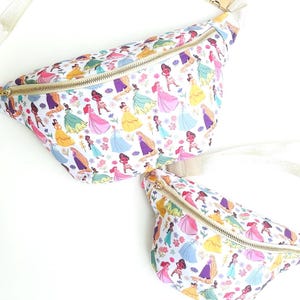 May include: Two white fanny packs with a colorful princess pattern. The bags feature a gold zipper and a beige strap. The pattern includes various princesses in their iconic dresses, along with floral accents.
