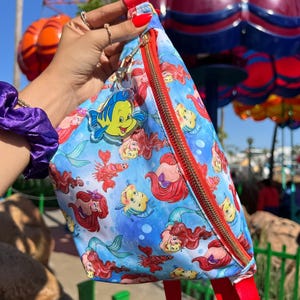 May include: A blue fanny pack featuring a repeating pattern of mermaids and fish. The bag has a red strap and zipper, with a gold-colored zipper pull. A Flounder charm hangs from the zipper. The background is a sunny outdoor scene.