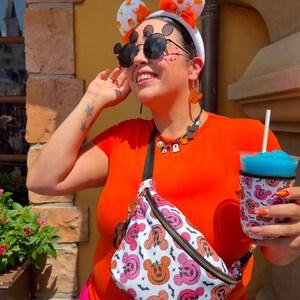 May include: A white headband with candy corn ears and an orange bow, black round sunglasses with mouse ear accents, a beaded necklace with character charms, and a white fanny pack featuring a pink and orange pumpkin mouse pattern with black bats.