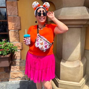 May include: A bright orange short-sleeved t-shirt, a vibrant pink tiered skirt, and a white fanny pack featuring a colorful Mickey Mouse-inspired Halloween pattern. The outfit includes white candy corn ears, round black sunglasses, and a matching drink sleeve.