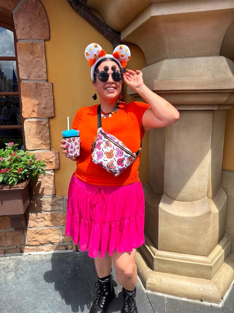 May include: A bright orange short-sleeved t-shirt, a vibrant pink tiered skirt, and a white fanny pack featuring a colorful Mickey Mouse-inspired Halloween pattern. The outfit includes white candy corn ears, round black sunglasses, and a matching drink sleeve.