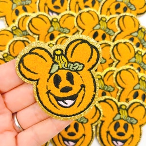 May include: Orange and gold glitter patches in the shape of a smiling pumpkin with mouse ears. The patches have a black outline and a green stem. The patches are a Halloween-themed craft supply.
