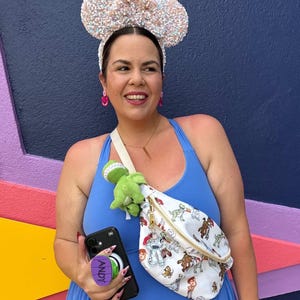 May include: A woman wearing a blue dress and sequined Mickey Mouse ears. She is holding a black phone with a purple "ANDY" pop socket and a white fanny pack with a Toy Story print. A green dinosaur plush is attached to the fanny pack.