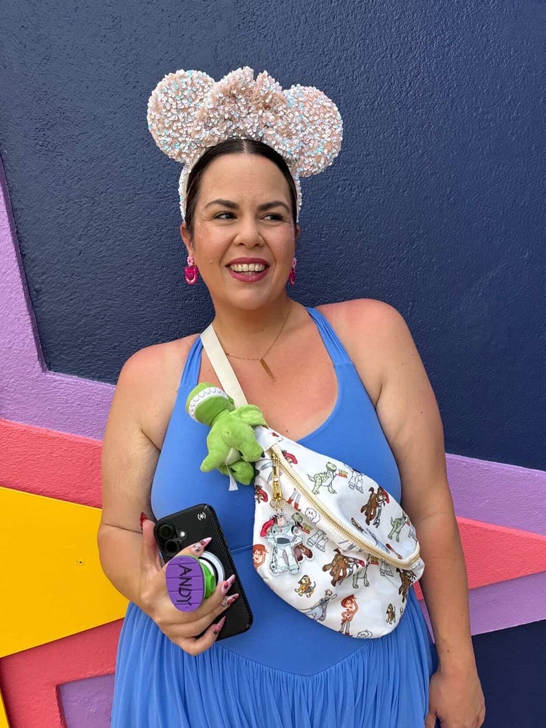 May include: A woman wearing a blue dress and sequined Mickey Mouse ears. She is holding a black phone with a purple "ANDY" pop socket and a white fanny pack with a Toy Story print. A green dinosaur plush is attached to the fanny pack.