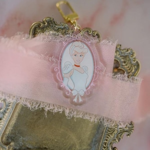 May include: A Cinderella-themed keychain with a gold-colored clasp. The keychain features a pink oval frame with a Cinderella illustration. The frame is set against a pink ribbon with a frayed edge, resting on a decorative gold-colored tray.