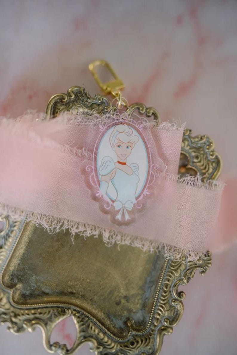 May include: A Cinderella-themed keychain with a gold-colored clasp. The keychain features a pink oval frame with a Cinderella illustration. The frame is set against a pink ribbon with a frayed edge, resting on a decorative gold-colored tray.